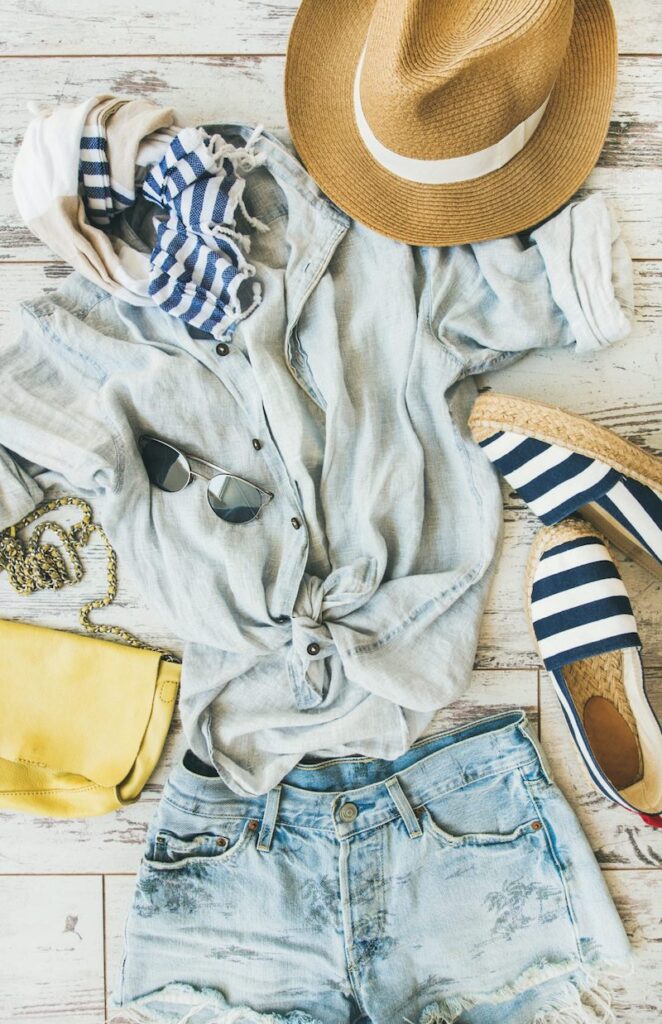 Pastel colored ethical clothing, parquet background, top view. Blue shorts, linen shirt, straw hat, yellow bag, sunglasses, striped neckerchief, espadrillas