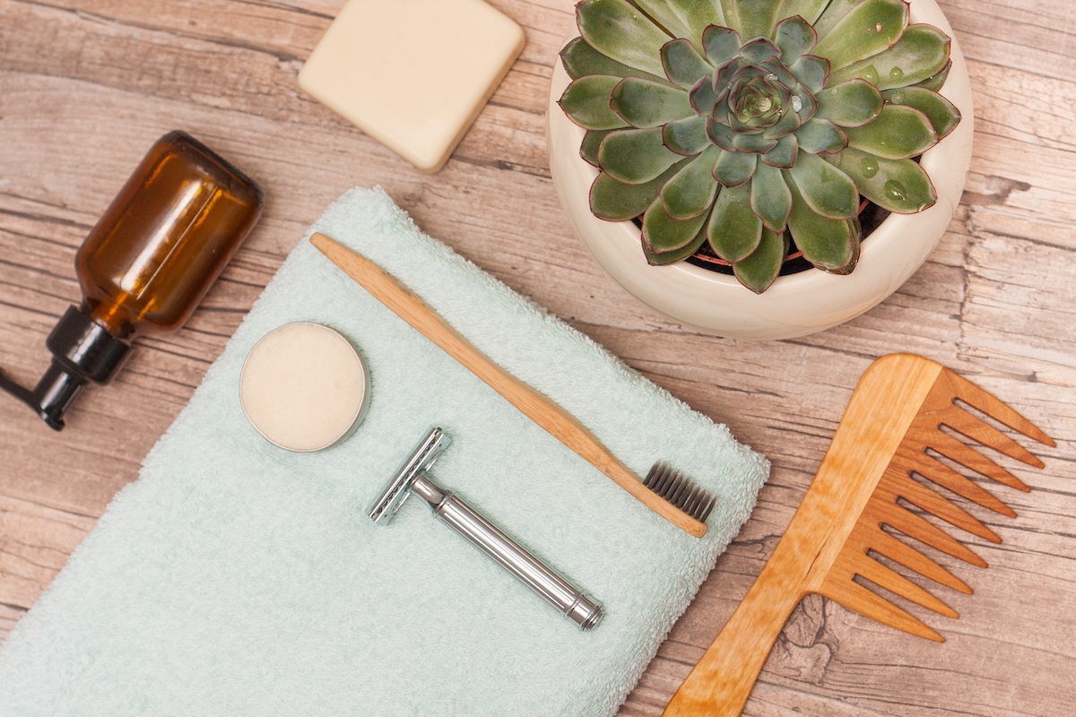 Zero waste bathroom accessories, metal safety razor, wooden comb, deodorant, shea butter, solid soap, wooden toothbrush, olive oil make up remover in a glass container, nail brush, aloe vera.